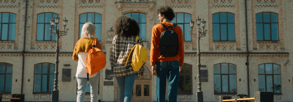 Three individuals with backpacks walk away from the camera towards a large, classical building with many windows and a clock tower.