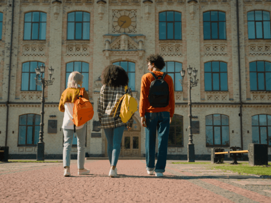 Three individuals with backpacks walk away from the camera towards a large, classical building with many windows and a clock tower.