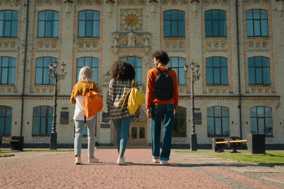 Three individuals with backpacks walk away from the camera towards a large, classical building with many windows and a clock tower.