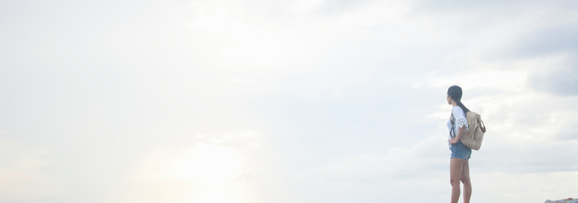 A young Asian woman hiker on vacation stands on a coastal rock with her backpack at sunrise and looks out to sea. Travel, wonderlust and achievement concepts.