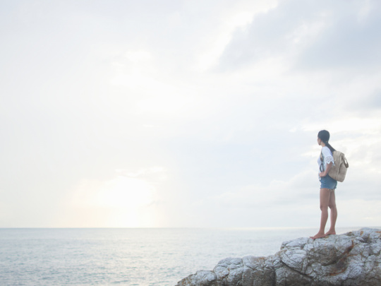 A young Asian woman hiker on vacation stands on a coastal rock with her backpack at sunrise and looks out to sea. Travel, wonderlust and achievement concepts.