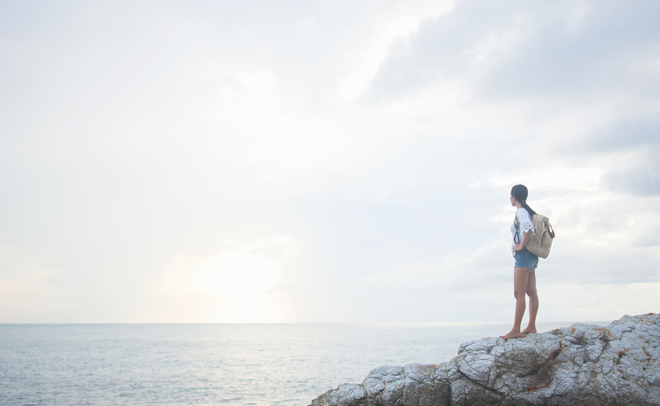 A young Asian woman hiker on vacation stands on a coastal rock with her backpack at sunrise and looks out to sea. Travel, wonderlust and achievement concepts.
